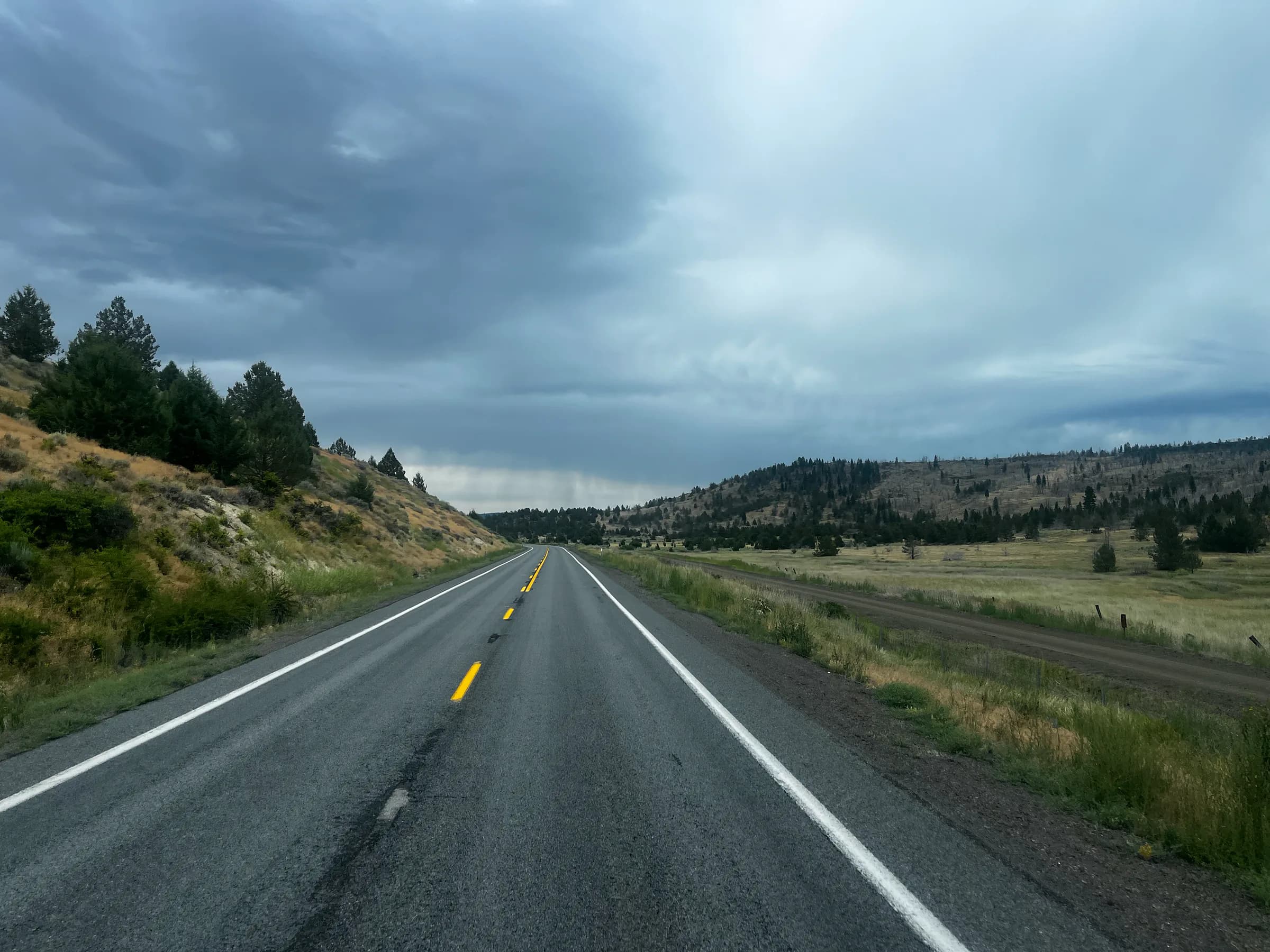A straight two-lane road running through meadow and pine hills toward rain falling from a dark sky.