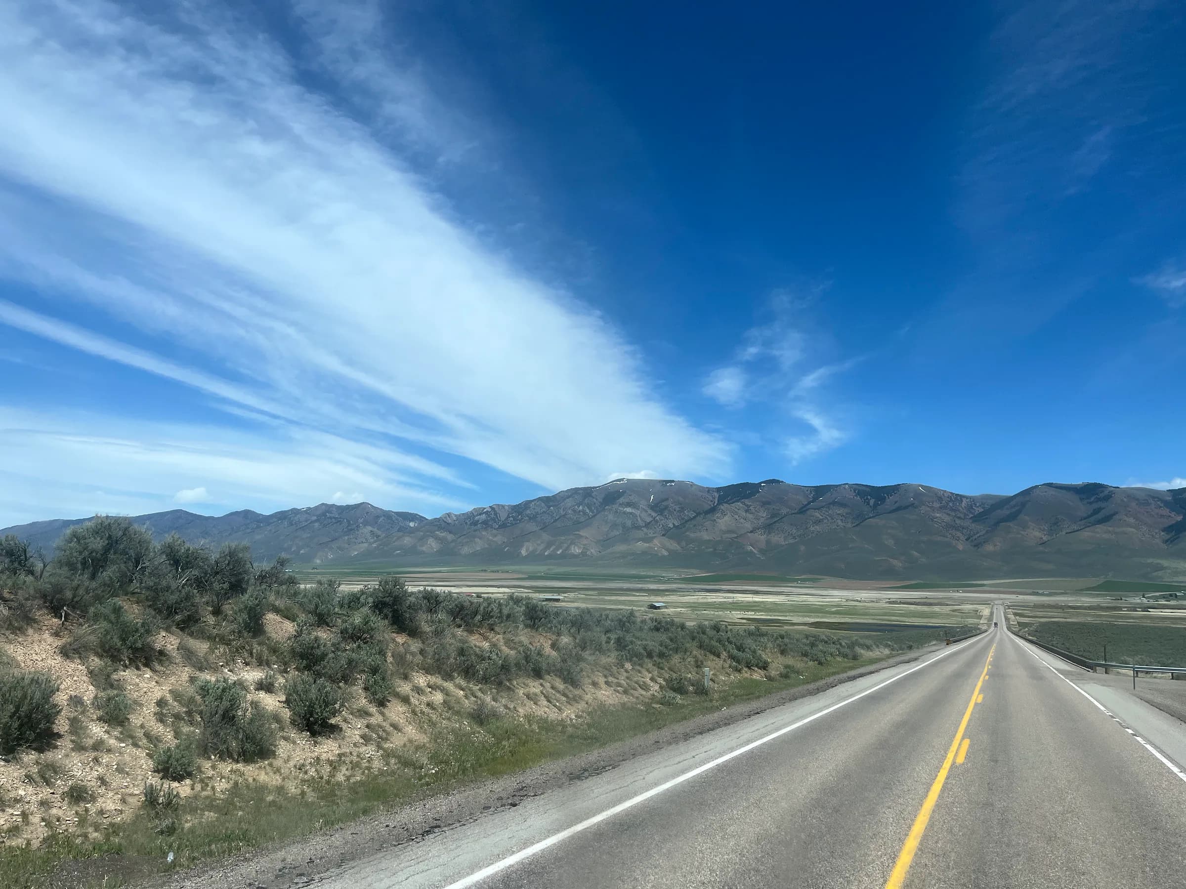 A highway running along sagebrush toward a long mountain range under sweeping streaks of cirrus.