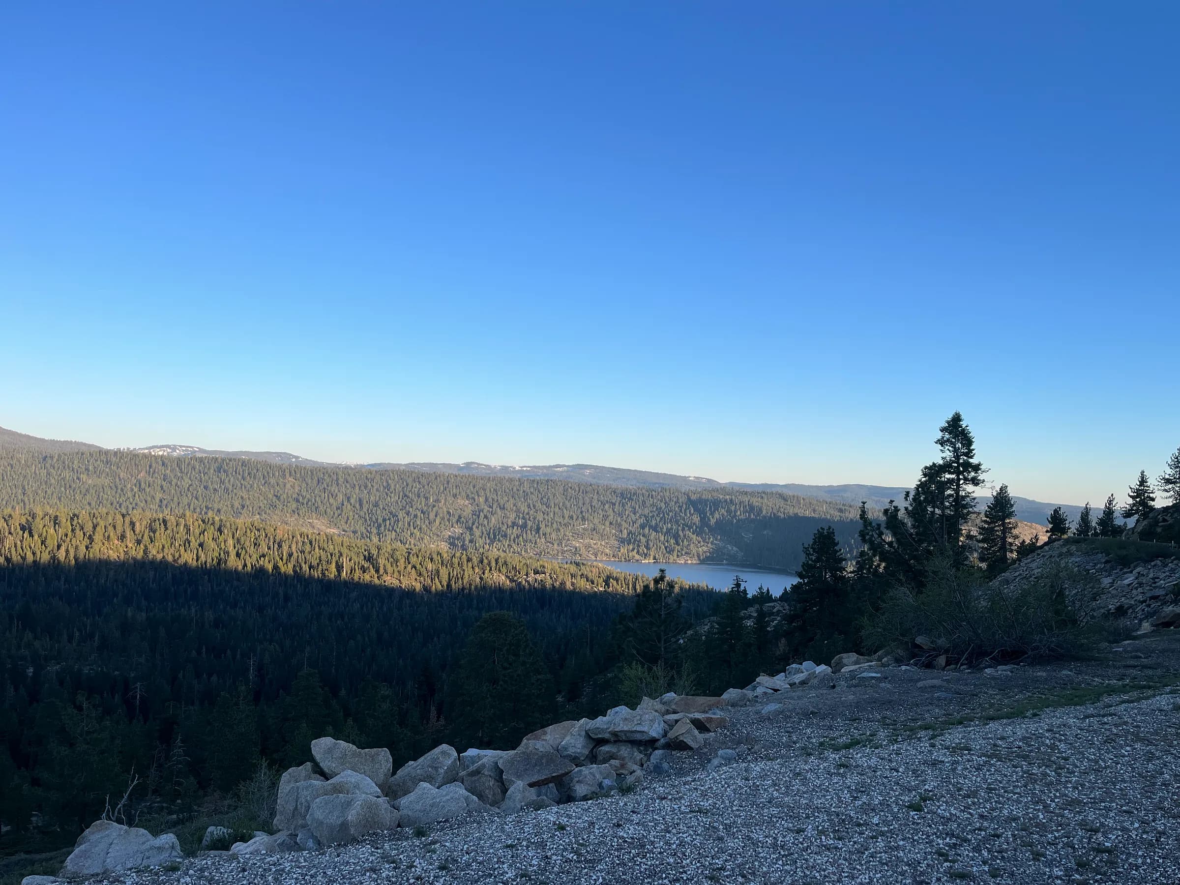 A forested valley seen from a gravel turnout high on a mountain road, a lake in the fold and pale ridges far off.