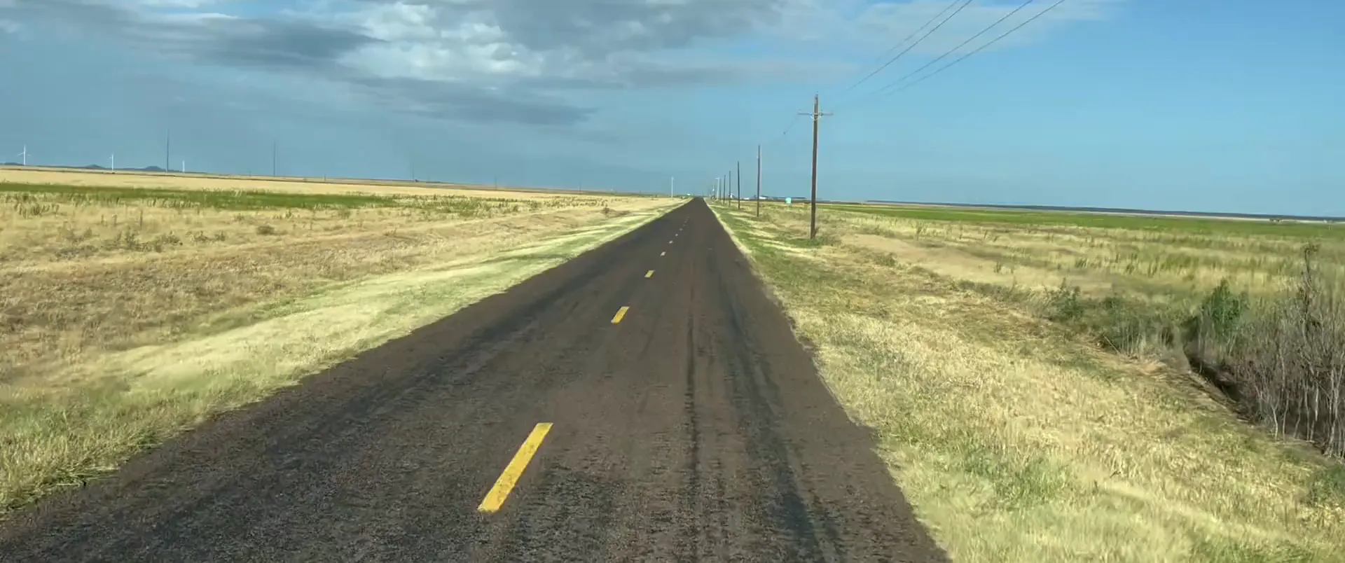Road footage: an empty two-lane farm road with yellow centre dashes running between wheat stubble on a clear morning.