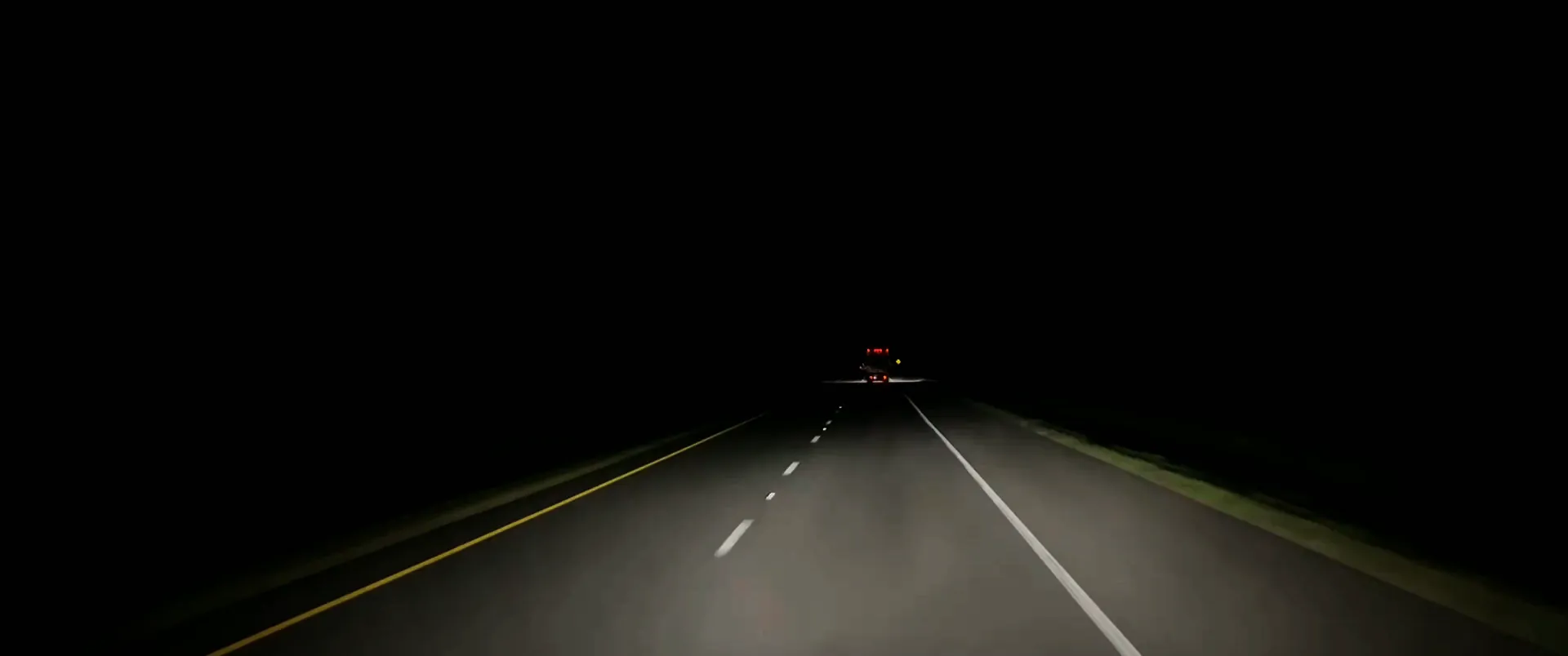 Road footage at night: an interstate lit by headlights, the red marker lights of a truck far ahead.